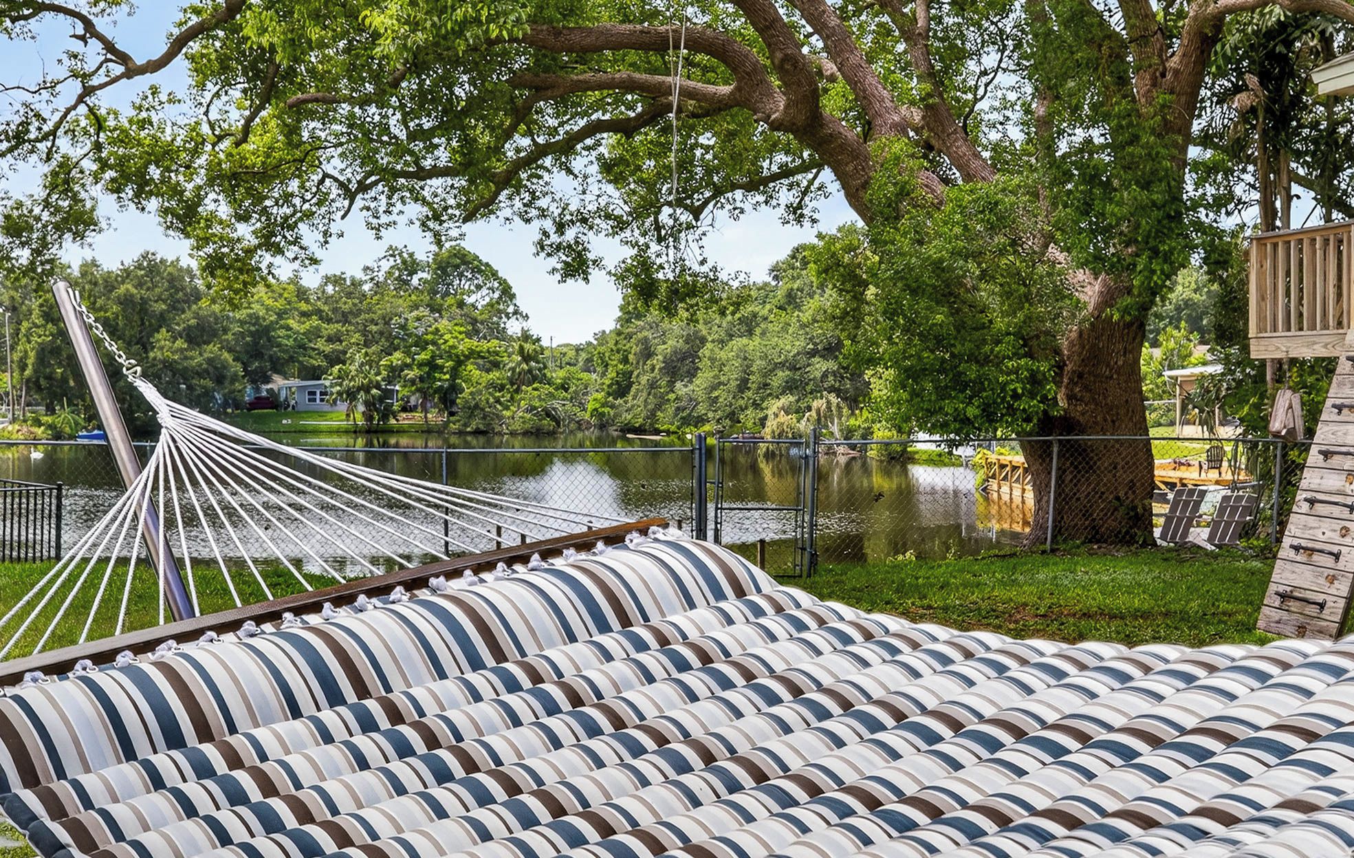 A hammock and striped cushions by a lake with trees and a fence in the background.