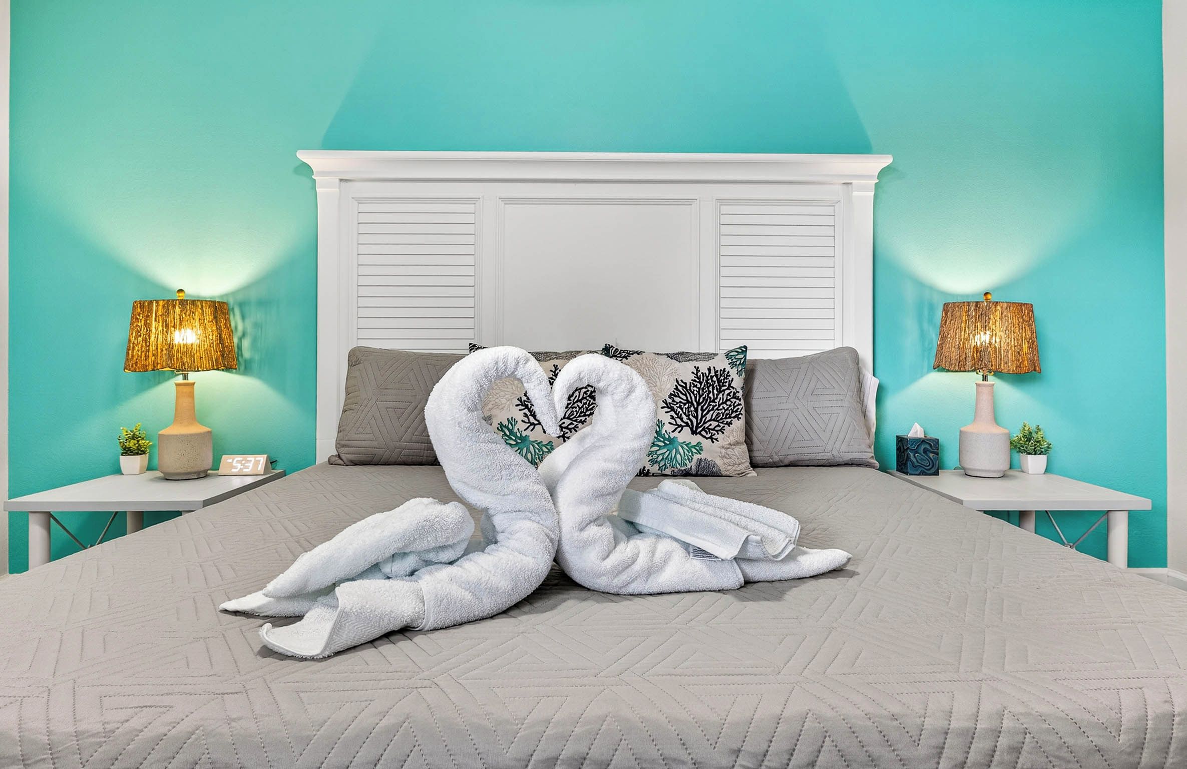 A bed with a white headboard, two nightstands with lamps, and a pair of towels arranged in a heart shape.