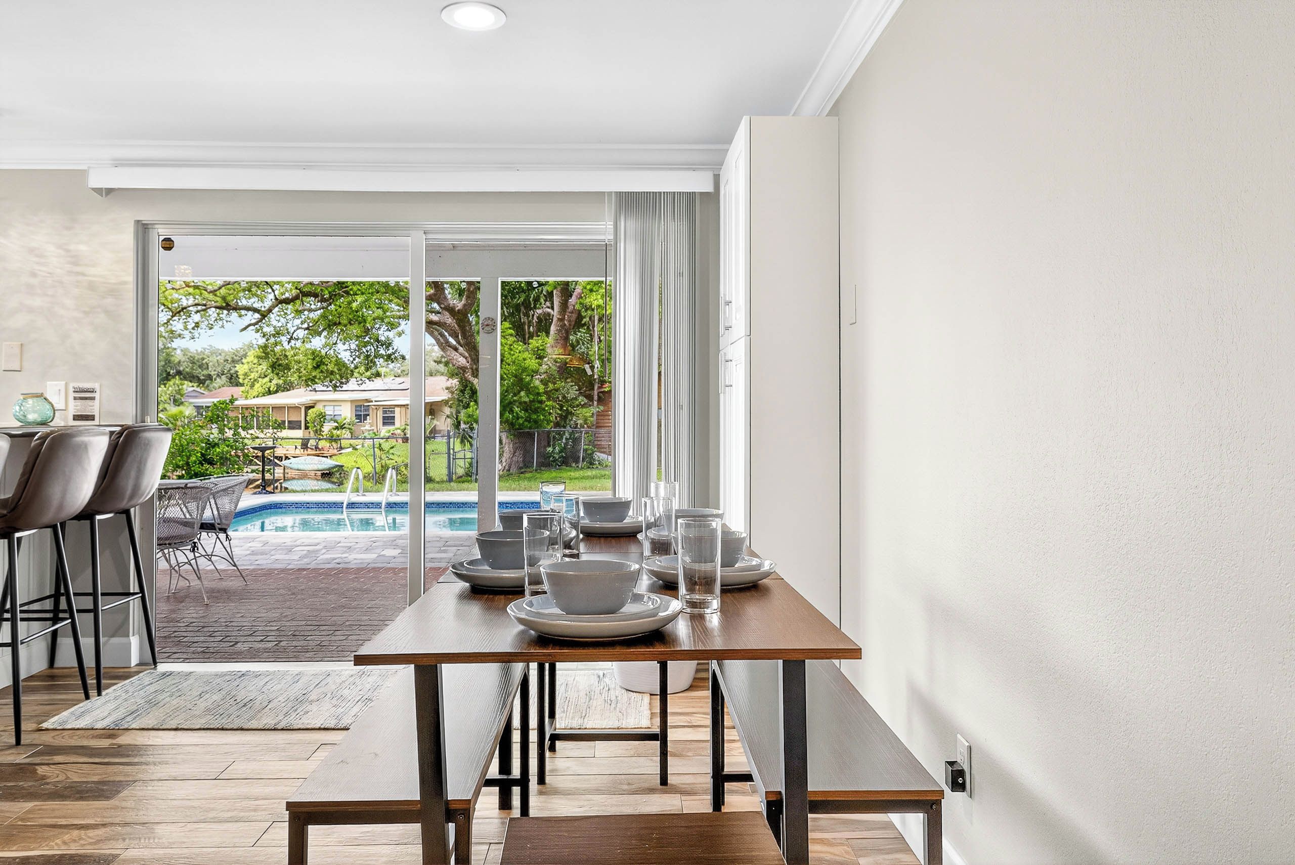 A dining area with a table set for four, a pool visible through the glass door, and a view of a residential area outside.