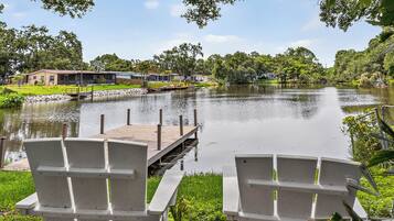 A lakeside view with a dock and two chairs facing the water, surrounded by greenery and a row of houses.