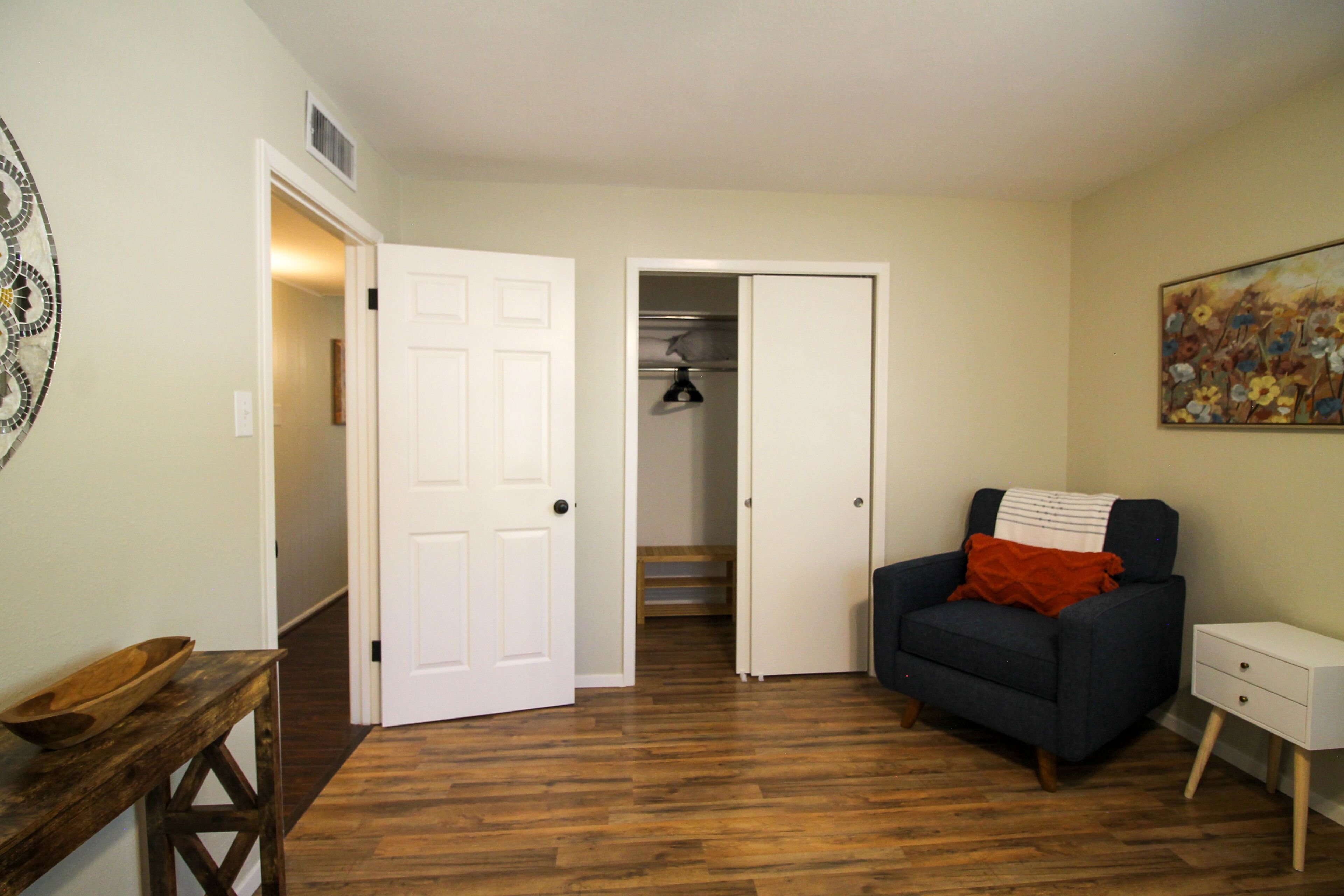 A room with a wooden floor, a dark blue armchair with a red pillow, a white bedside table, and a door leading to a closet with shelves.