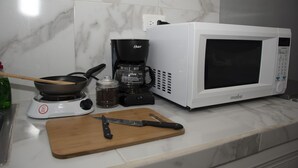 Eine Küche mit einer weißen Mikrowelle, einer Kaffeemaschine, einer Pfanne, einem Holzer Schneidebrett und einem Messer.