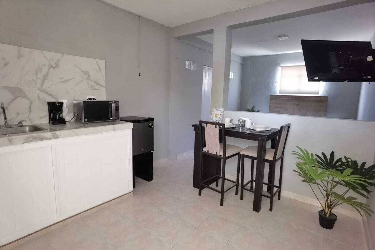 A modern kitchen with a white countertop, a black microwave, and a coffee maker. There is a dining table with chairs and a potted plant.