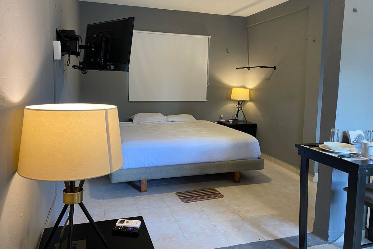 A hotel room with a bed, bedside table, lamp, TV, and a small table with a cup and saucer.