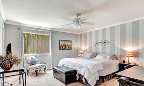 Une chambre à coucher comprenant un lit, un bureau, une chaise, une fenêtre avec des stores, un ventilateur de plafond et un tableau accroché au mur.