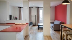 A modern kitchen with a red countertop, a dining area with a table and chairs, and a living area with a red wall and a sofa. - Apartamento Premium 2 Dormitorios 1 Baño con Balcón (Madrid)