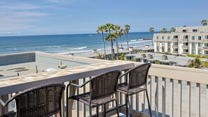 A balcony with wicker chairs overlooking the ocean, a building with balconies, and palm trees. - Top Floor Panoramic Whitewater Ocean Views in Oceanside! 1,081 Sq Ft (Oceanside)