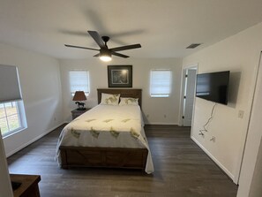 A bedroom with a bed, ceiling fan, TV, and two windows.