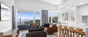 A modern living room with a sofa, a dining area with bar stools, and large windows offering a view of the sea.