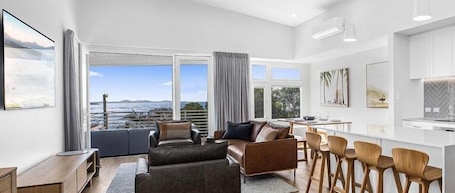 A modern living room with a sofa, a dining area with bar stools, and large windows offering a view of the sea.