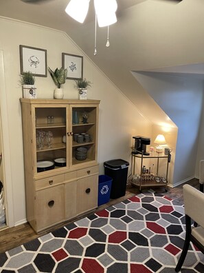 Un rincón de una habitación con un armario de madera, un bote de reciclaje azul, una cafetera y una alfombra estampada.