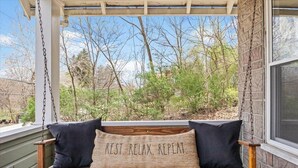 A wooden swing with black cushions and a pillow with the text "REST. RELAX. REPEAT." in a covered outdoor area with a view of trees and a building in the background. - Family and dog friendly☆3BR house☆Porch swing☆Mars☆PGH☆Cranberry (Mars)