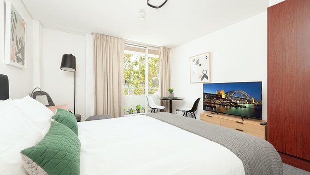 A hotel room with a large bed, a TV displaying a cityscape, a desk with a chair, and a window with curtains. - Cremorne - MP303 Military Road - Convenient Studio in the Heart of Cremorne with Shops, Dining Transport at Doorstep (Cremorne)