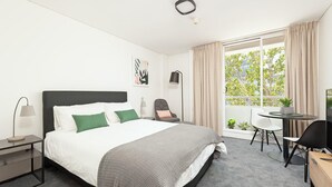 A modern hotel room with a bed, bedside tables, a desk with a chair, a window with curtains, and a small balcony. - Cremorne - MP303 Military Road - Convenient Studio in the Heart of Cremorne with Shops, Dining Transport at Doorstep (Cremorne)