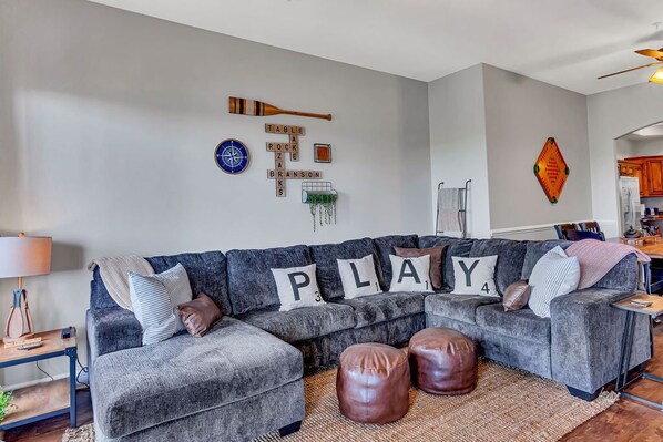 Un salon moderne avec un canapé d’angle gris, des poufs en cuir et une décoration murale composée de mots et d’une boussole.