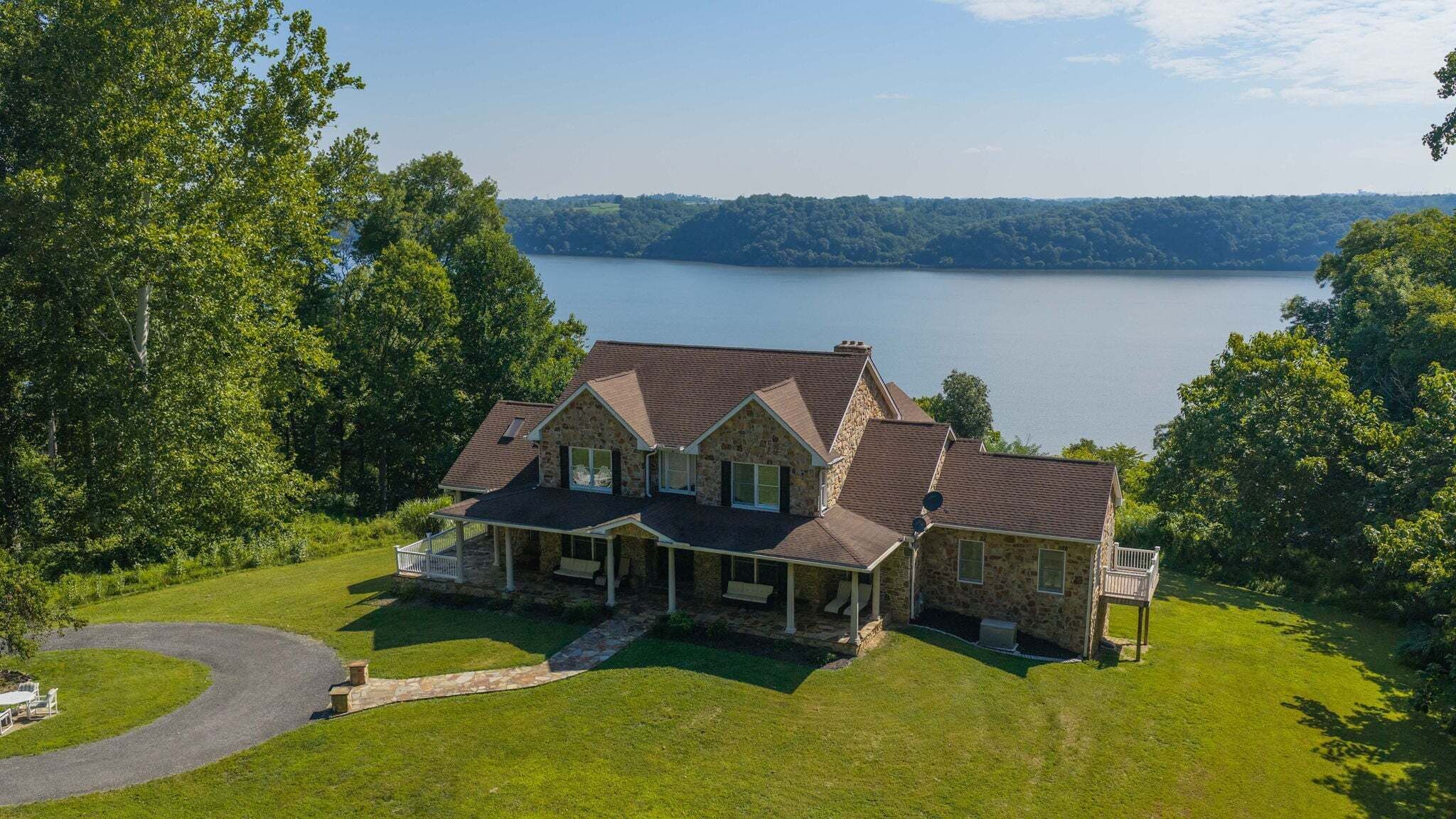 A two-story stone house with a wrap-around porch, a stone chimney, and a view of a lake.