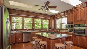 Una cocina con armarios de madera, una isla grande con taburetes, un ventilador de techo y vistas amplias a través de grandes ventanales.