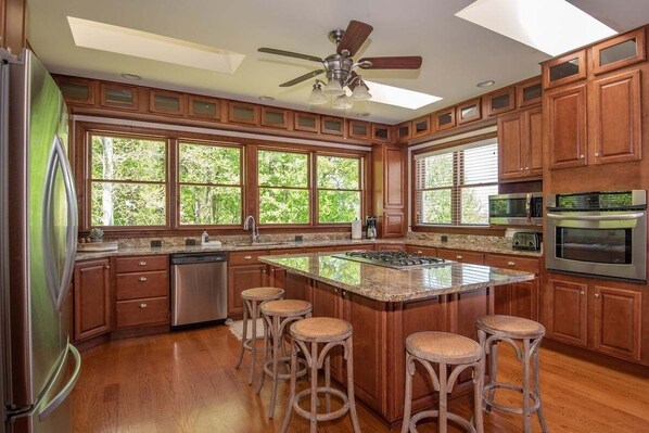 Una cucina con mobili in legno, una grande isola con sgabelli, una ventola a soffitto e vista panoramica attraverso ampie finestre.