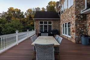 A deck with a wooden table and wicker chairs, a stone building with a chimney, and a white railing. - Waterfront Retreat w/ Hot Tub, Games & Views (Whiteford)