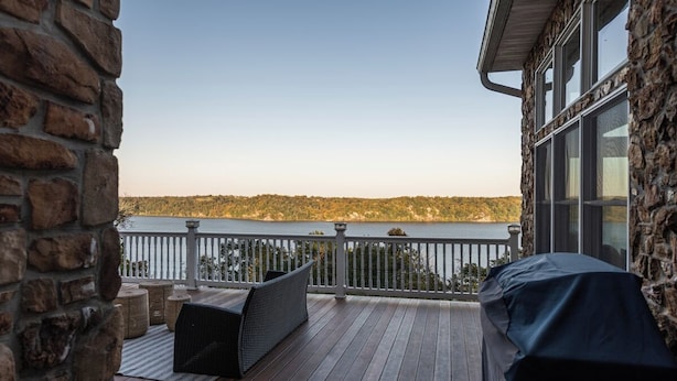 Una terraza de madera con un muro de piedra, una parrilla cubierta y vista a un lago.