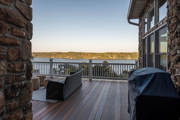 Eine Holzterrasse mit einer Steinmauer, einer Grillstelle unter Vordach und Blick auf einen See.