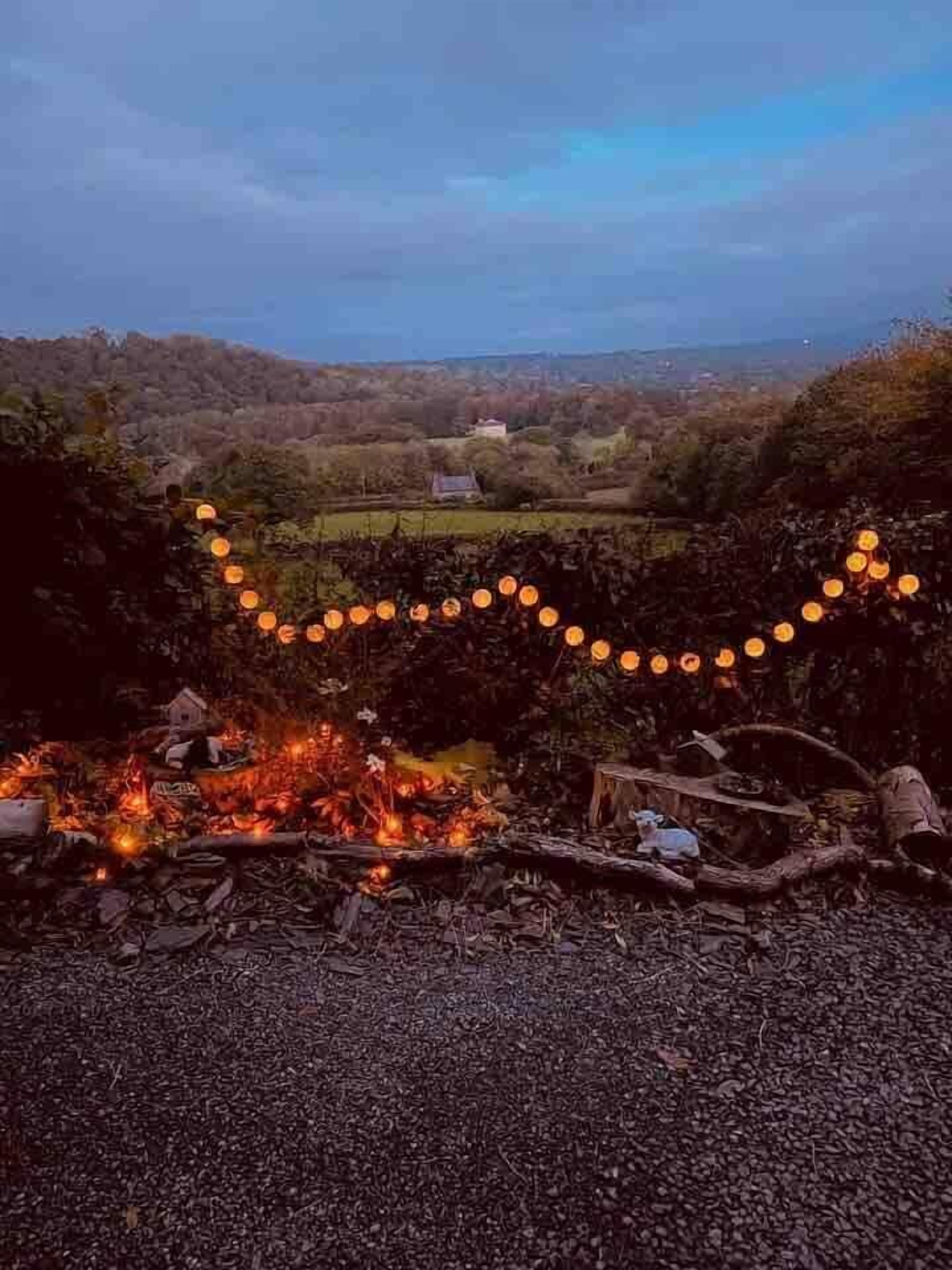 Un campament amb foc, envoltat d'arbres i vista cap a una casa llunyana.
