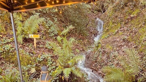An outdoor seating area with a table, cushions, and a backdrop of a waterfall and foliage. - A beautiful, unique lodge, made for two (Lampeter)