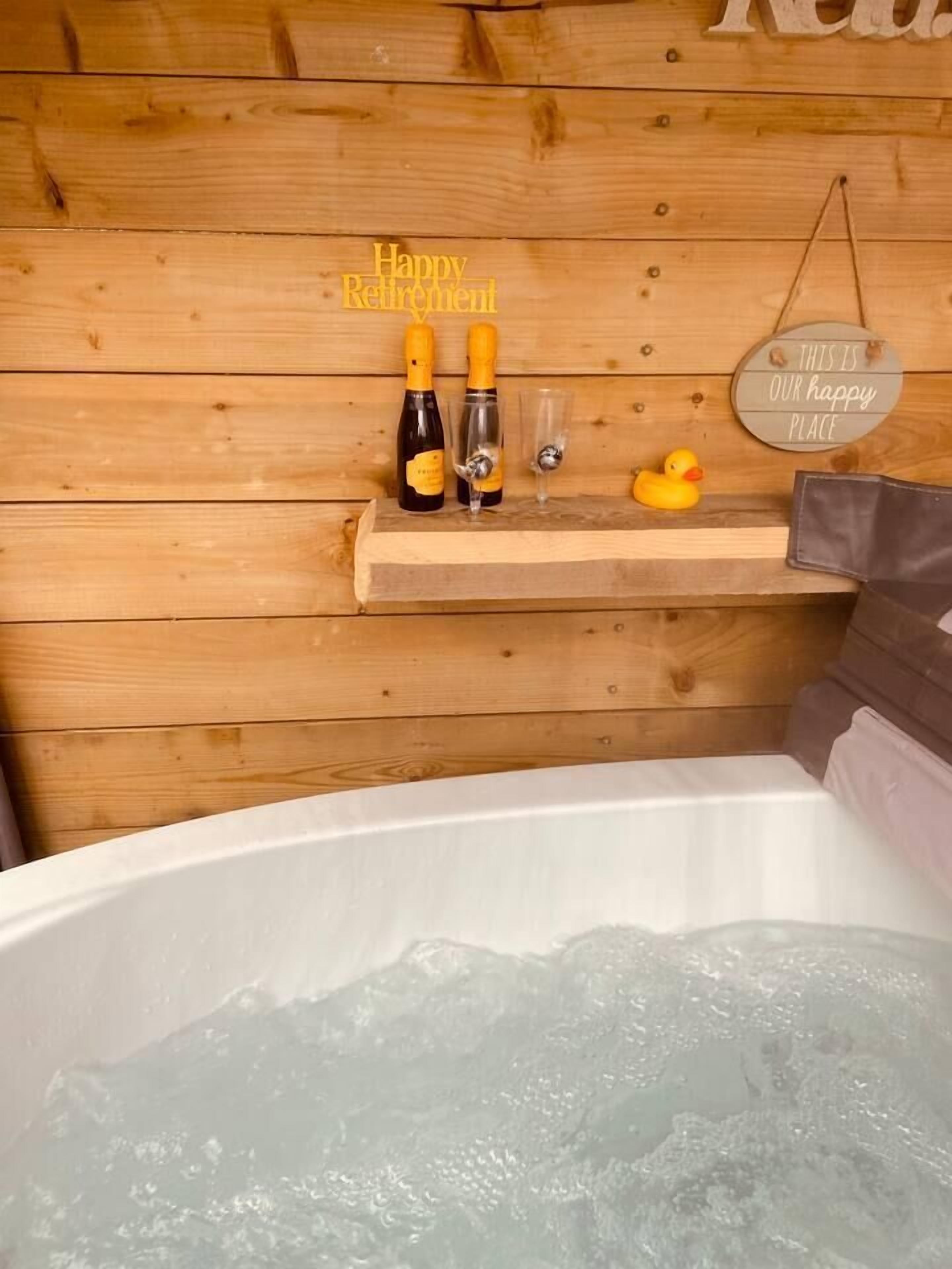 A wooden cabin interior with a hot tub, two champagne bottles, glasses, and a rubber duck. A sign reads "Happy Retirement."