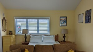 Een woonkamer met een bruine bank, twee lampen, een bed met witte lakens, een raam met zonwering en een gele wand.