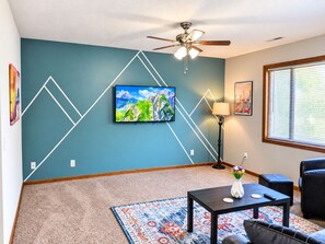 Ein Wohnzimmer mit einer türkisfarbenen Akzentwand, einem Wand-Fernseher, einem Deckenventilator, einer Lampe, einem Couchtisch, einer Couch und einem Fenster mit Jalousien.