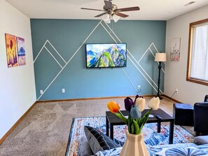 Un salon avec un téléviseur fixé au mur, un ventilateur de plafond, une table ornée d’un vase de fleurs et un tapis à motifs.
