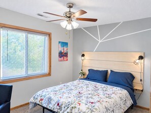 Une chambre à coucher comprenant un lit, un ventilateur de plafond, une fenêtre avec des stores et un mur orné d’un motif géométrique.