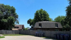 A parking lot with a grassy area, a wooden fence, and a building with a dark roof.