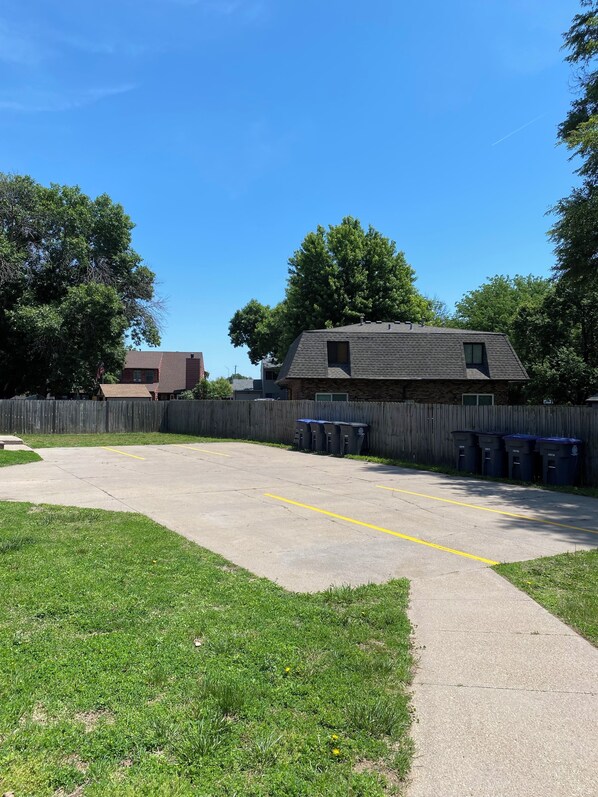 A parking lot with a grassy area, a wooden fence, and a building with a dark roof. - Renovated 2BR Retreat • Minutes from Downtown Omaha (Council Bluffs)