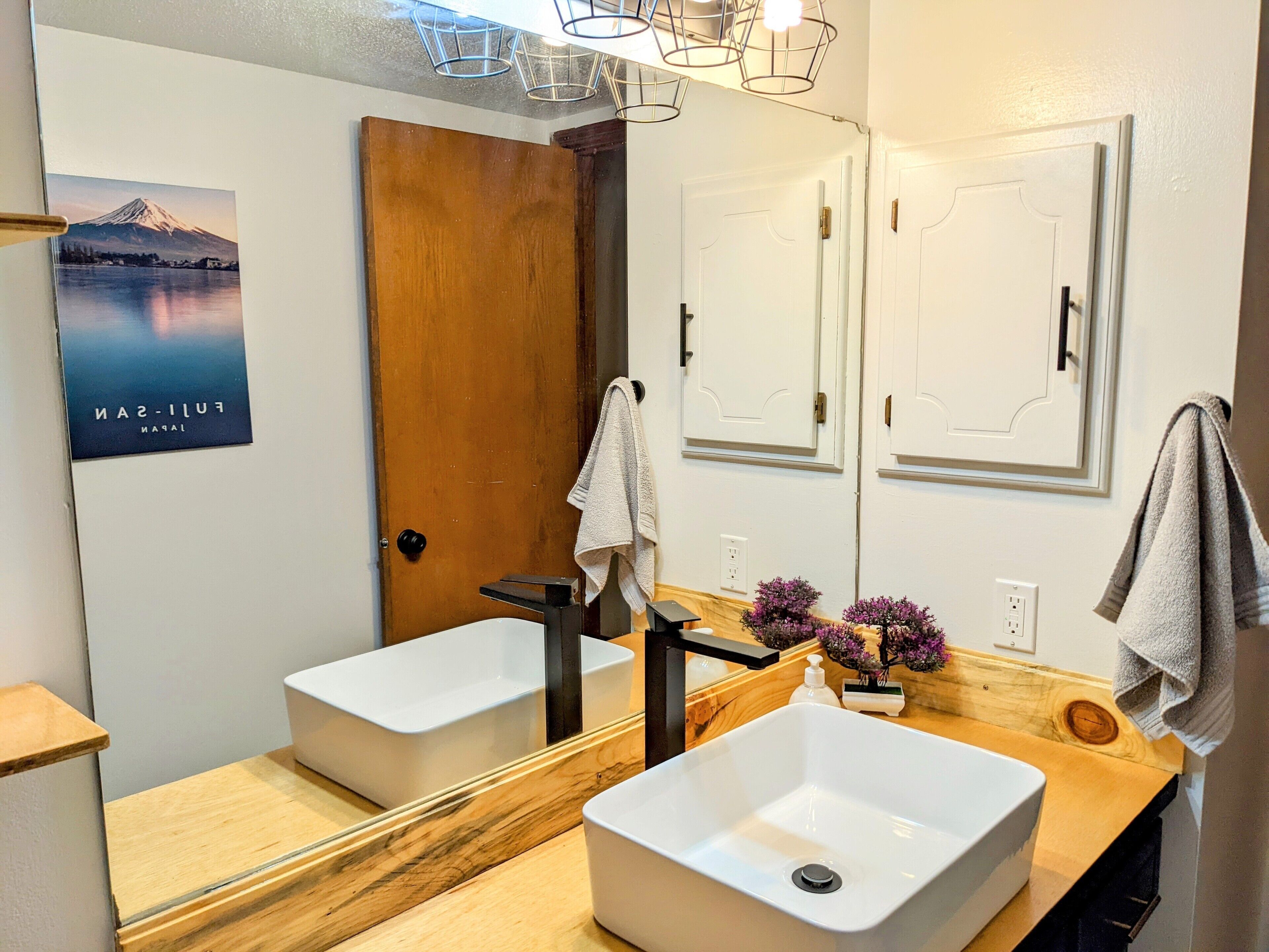 A modern bathroom with two sinks, a wooden vanity, a large mirror, and a door with a towel hanging on it.