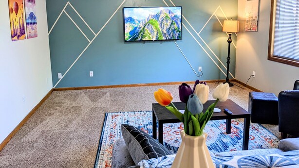 A living room with a ceiling fan, a mounted TV displaying a mountain landscape, a floor lamp, and a vase with flowers on a coffee table.