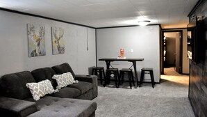 Una habitación con un sofá gris, dos cuadros en la pared, una mesa con taburetes y una puerta que da a otra estancia.
