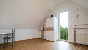 A room with a dining table, chairs, a white cabinet with a plant, a clock on the wall, and a view through a large window. - Alpenblick Apartment with Mountain View, Balcony, and Wi-Fi (Friedrichshafen)