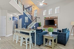 Un vaste espace de vie comprenant un escalier, une table à manger avec des tabourets, un canapé bleu et un téléviseur fixé au mur.
