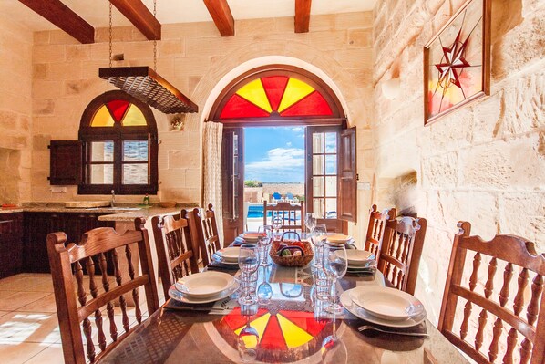 Un coin repas avec une table dressée pour six personnes, des chaises en bois, un mur en pierre et une vue dégagée sur l’extérieur grâce à une arche ouverte.