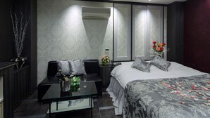 Une chambre d’hôtel moderne avec un grand lit, un coin salon doté d’une table en verre et une table de chevet agrémentée d’un vase de fleurs.