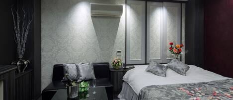 A modern hotel room with a large bed, a sitting area with a glass table, and a nightstand with a vase of flowers.