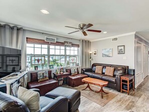 A living room with a leather sofa, a wooden coffee table, and a TV. There are windows with curtains, a ceiling fan, and a sign that says "Beach Rules". - Tybee South Beach Condo, Heated Pool Access (Tybee Island)