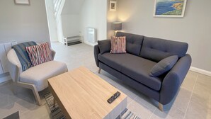Una sala moderna con un sofá gris, una mesa de centro de madera y un sillón a rayas. Se observa un radiador, una lámpara y un cuadro en la pared.