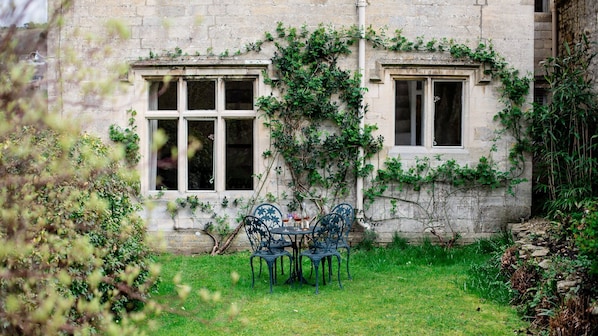 Relax in a serene outdoor seating area with a metal table and four chairs, surrounded by lush greenery and a traditional stone building.