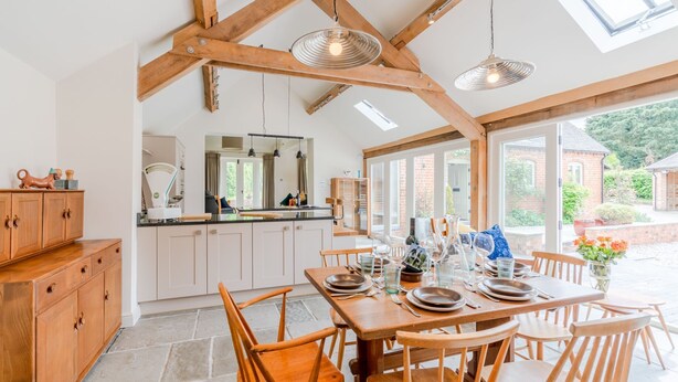 A spacious kitchen with wooden beams, a dining area with a table set for four, and a view of a brick building and greenery outside.