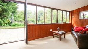 A room with a large sliding glass door leading to a garden, a wooden wall, a leather sofa with red cushions, and a small wooden table with a decorative item. - The Hermitage (Holt)