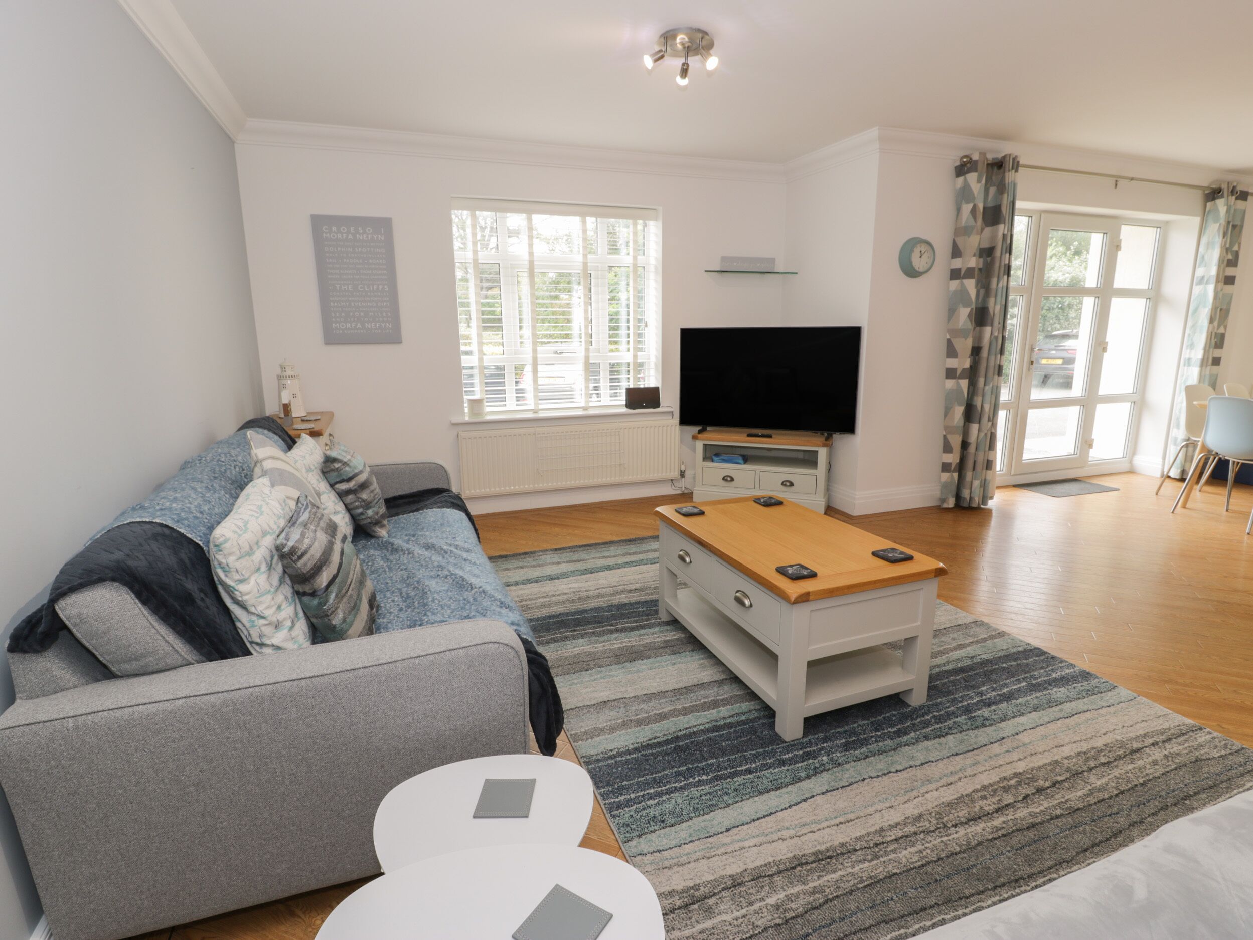 A living room with a grey sofa, a wooden coffee table, a flat-screen TV, and a large window with curtains.