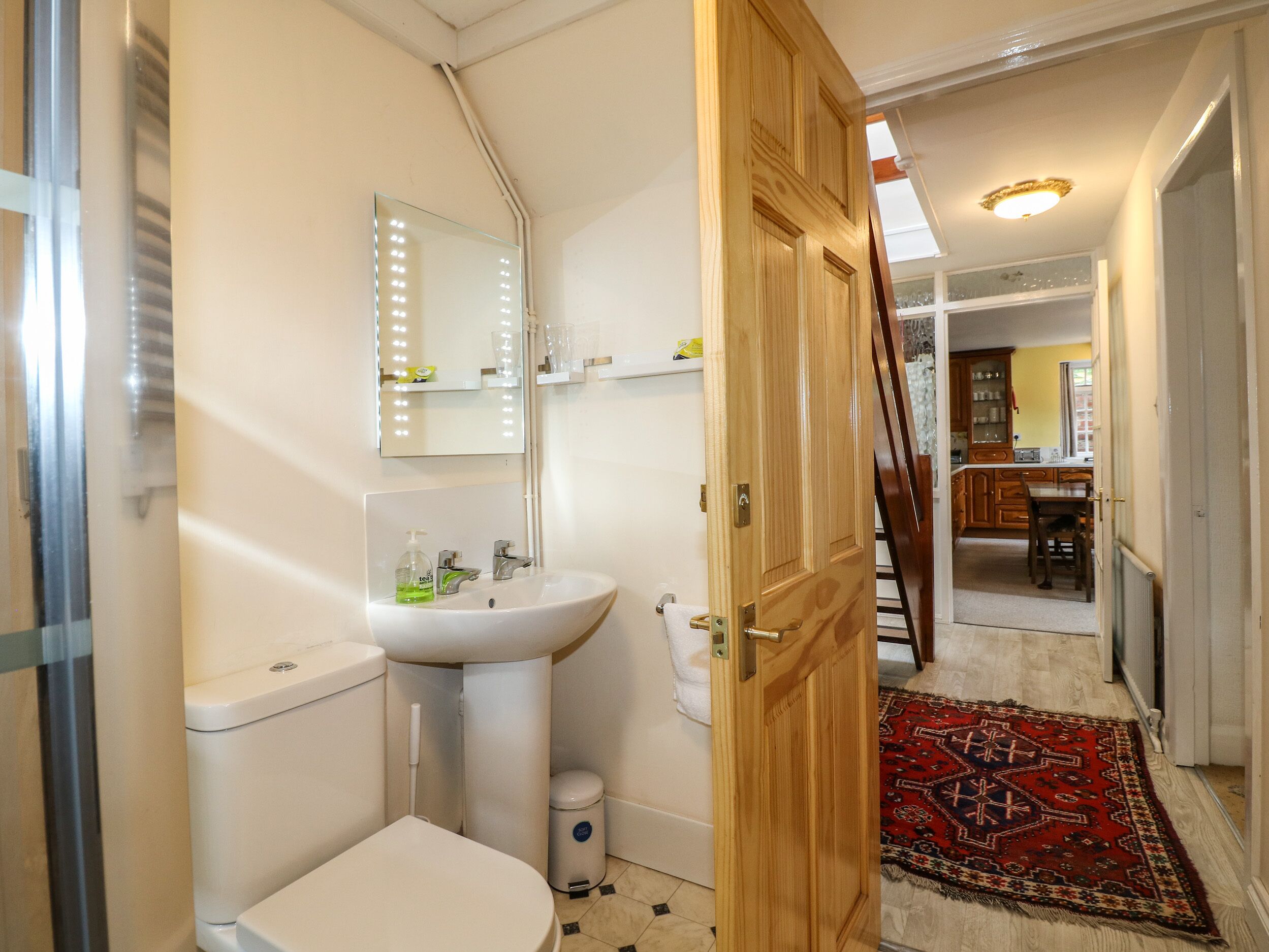 Baño con lavamanos, inodoro y espejo con iluminación. Se ve una puerta de madera que da a un pasillo con alfombra y escalera.