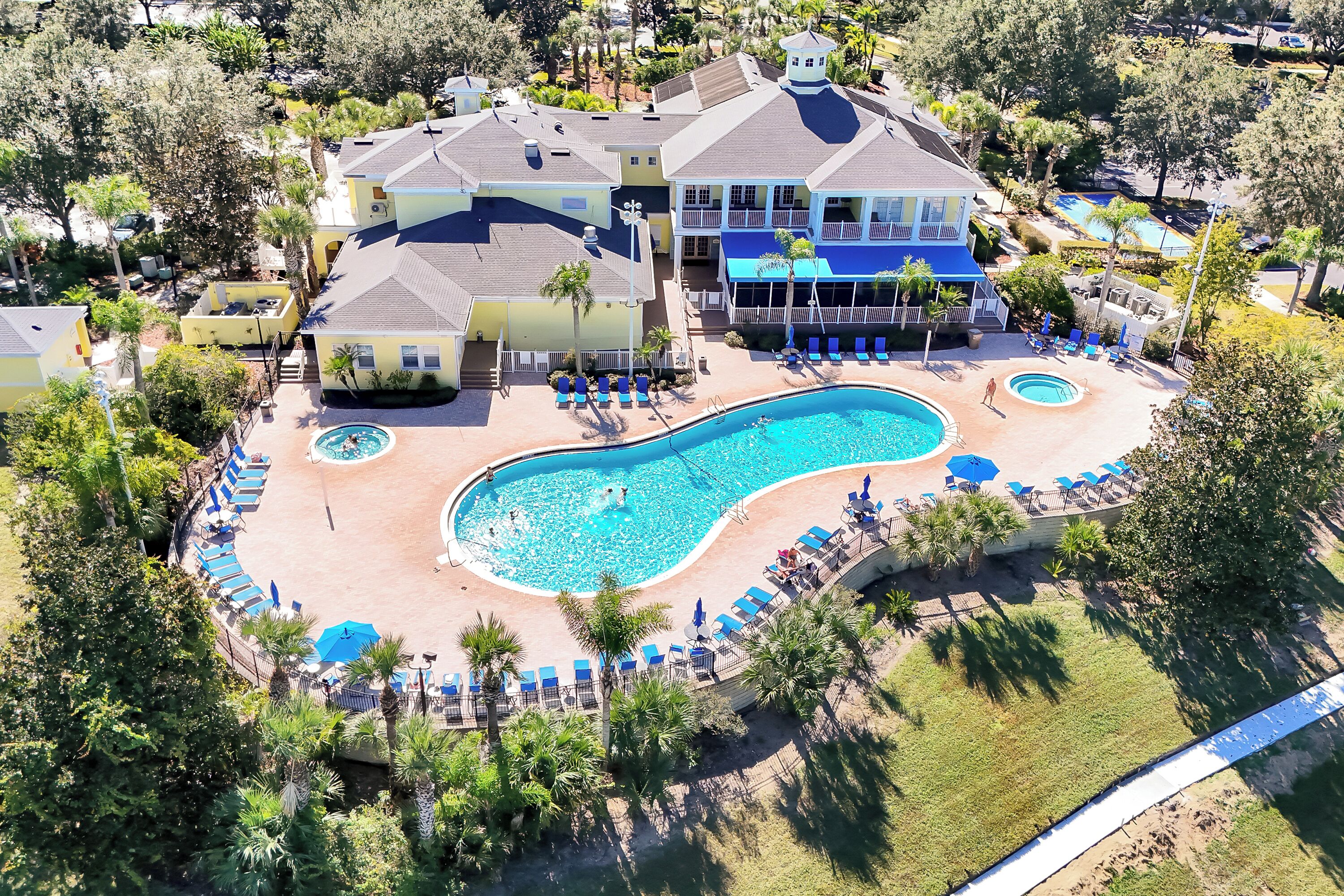 An aerial view of a resort with a large swimming pool, multiple lounge areas, and a variety of trees and greenery.